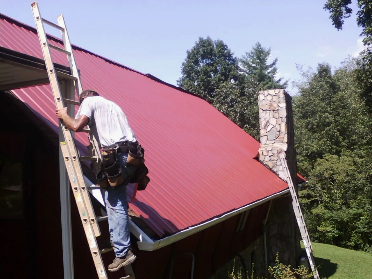 Roofer on a ladder beside a red metal roof and stone chimney — Soffit & Fascia Repair by Roofing Brothers in Prinsburg